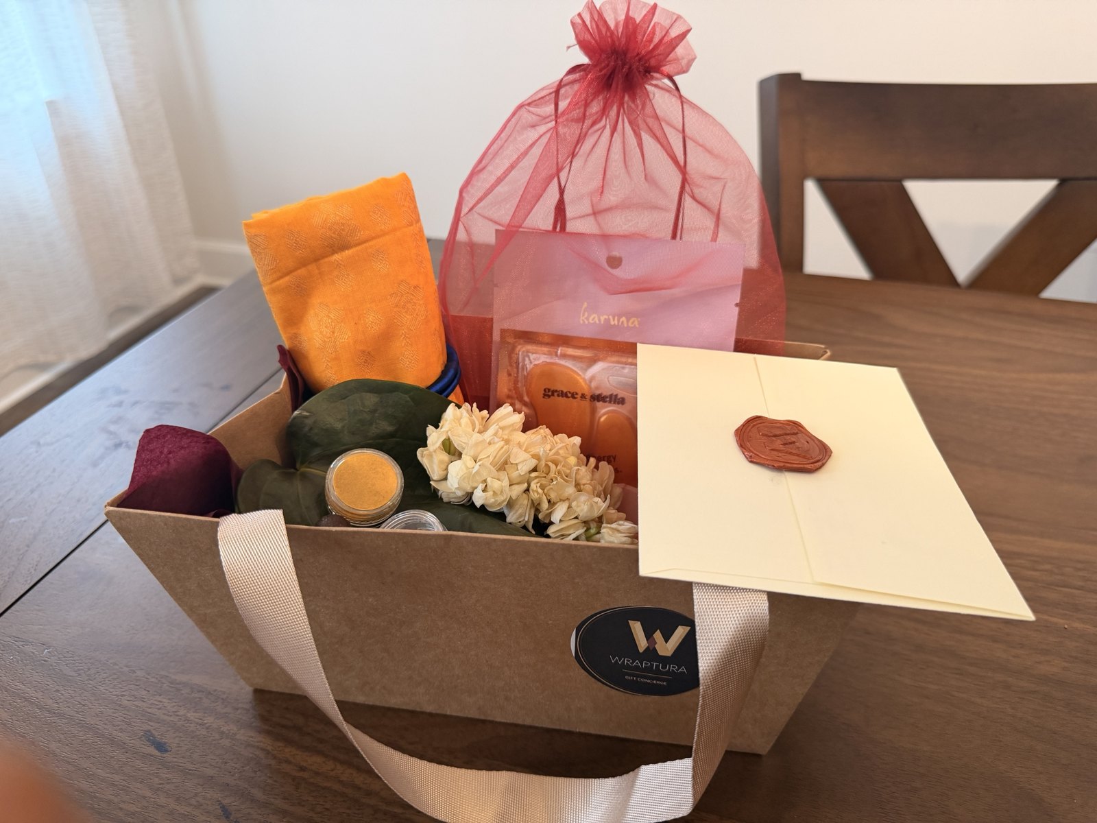 A finished Wraptura thamboolam tray in daylight: kraft carrier with satin handles, gold silk blouse piece, betel leaves, haldi and kumkum jars, fresh jasmine, an organza pouch of skincare, and a wax-sealed cream envelope